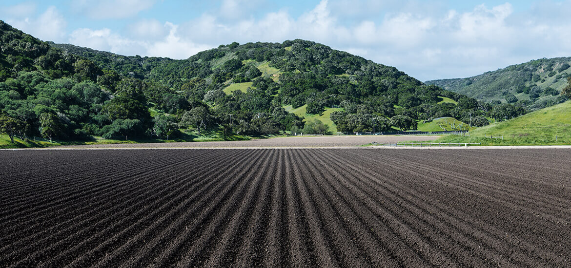 Picture of a freshly plowed farm field with rich black soil in neat rows surrounded by wooded hills.