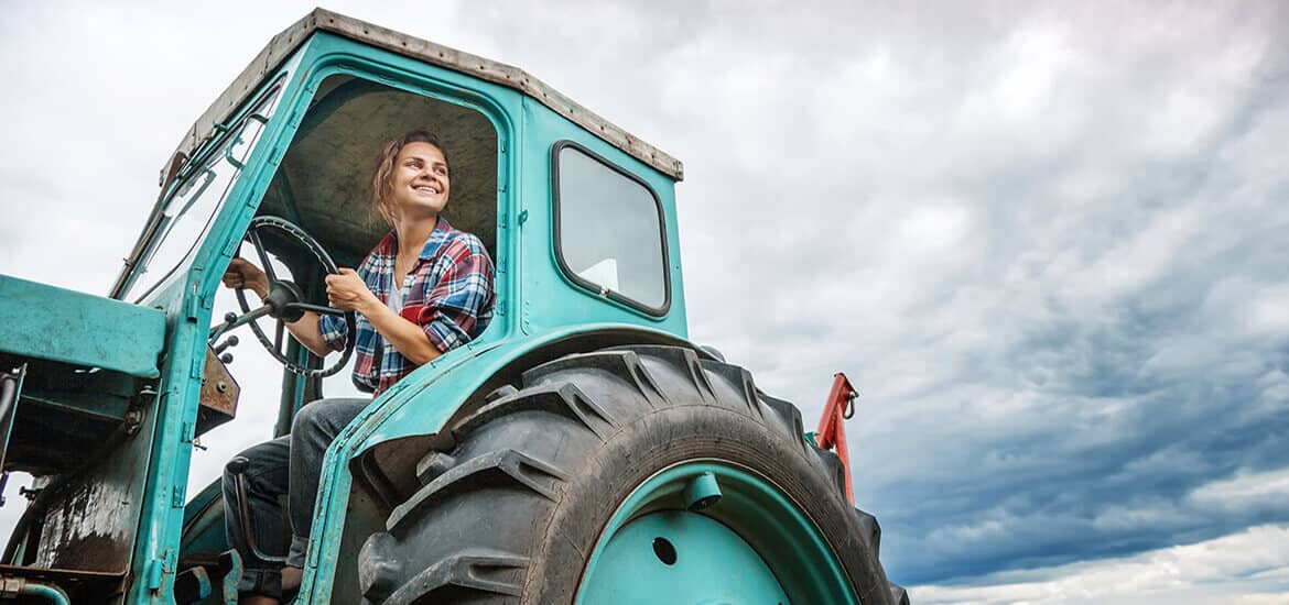 Picture of young female farmer in plaid shirt seated in the driver's seat of a vintage powder blue farm tractor.