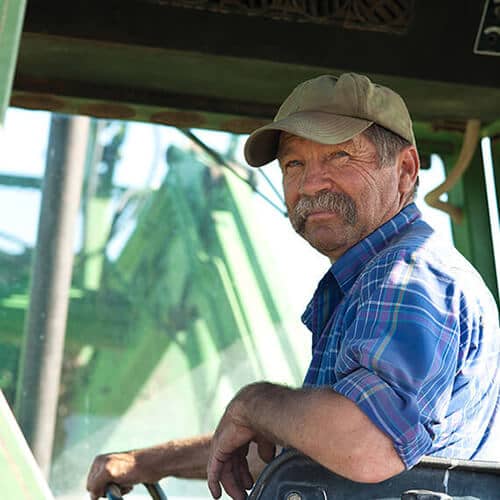 Picture of smiling farmer with moustache wearing plaid shirt riding in tractor cab.
