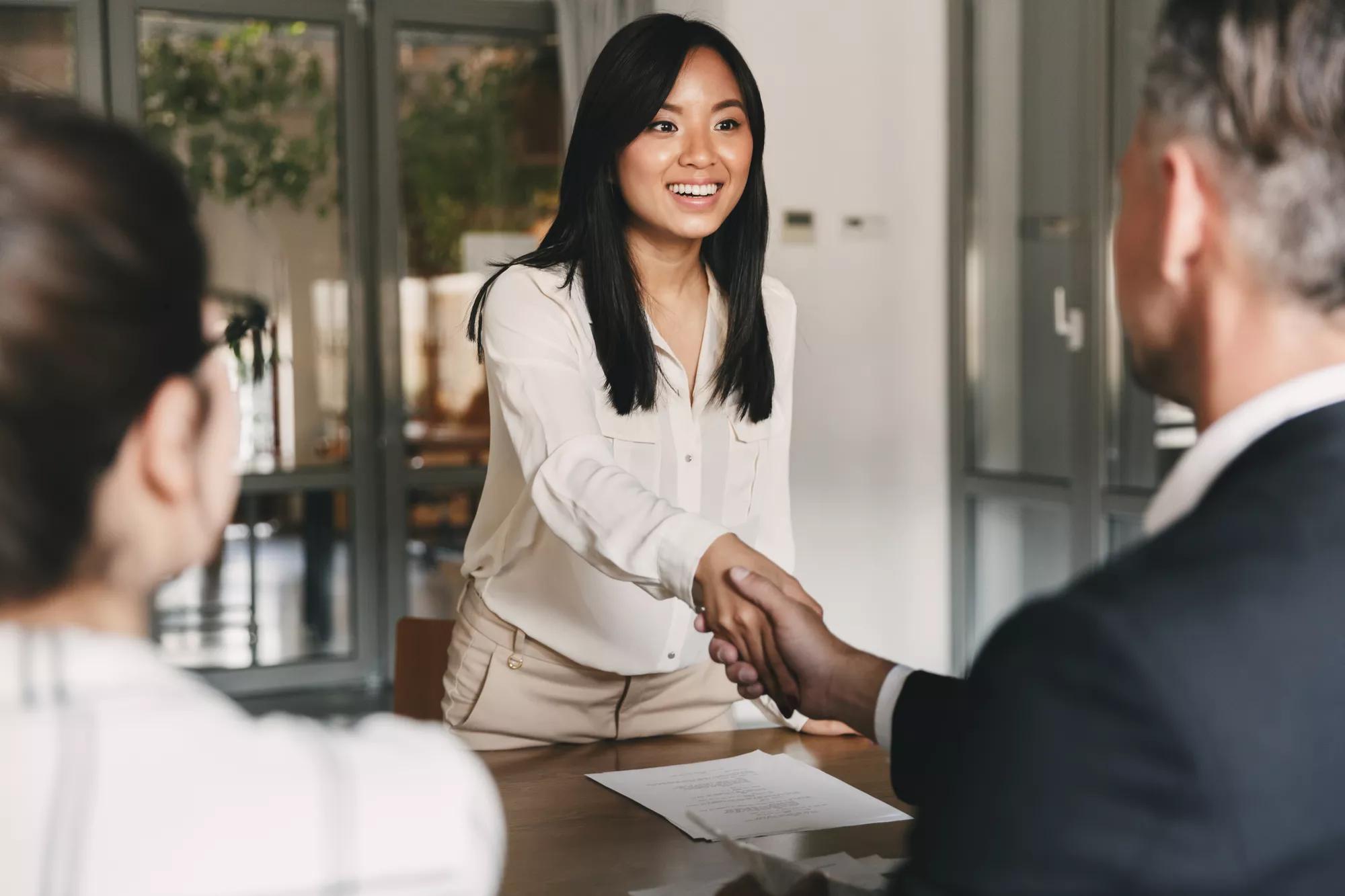 Back of two employers sitting in office and shaking hand of young asian woman, after successful negotiations or interview