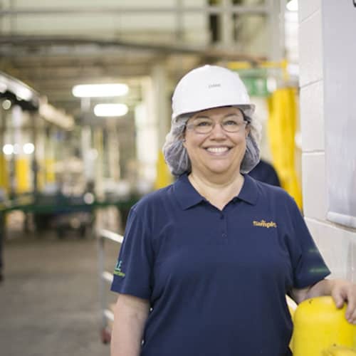 Picture of smiling woman wearing Simplot blue uniform and white bump helmet inside J.R. Simplot Company food processing plant.